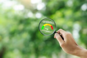 hand holds a magnifying glass in front of a window looking out at trees. On the magnifying glass is a meter showing good indoor air quality.